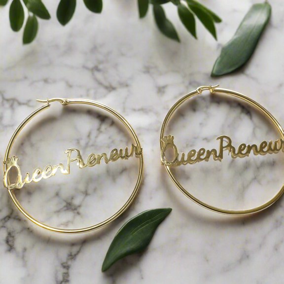 A pair of large, elegant gold hoop earrings featuring the word “QueenPreneur” in a bold cursive script, with a delicate crown accent over the “Q.” The earrings have a secure latch-back closure and a stylish, empowering design perfect for entrepreneurs and boss queens.
