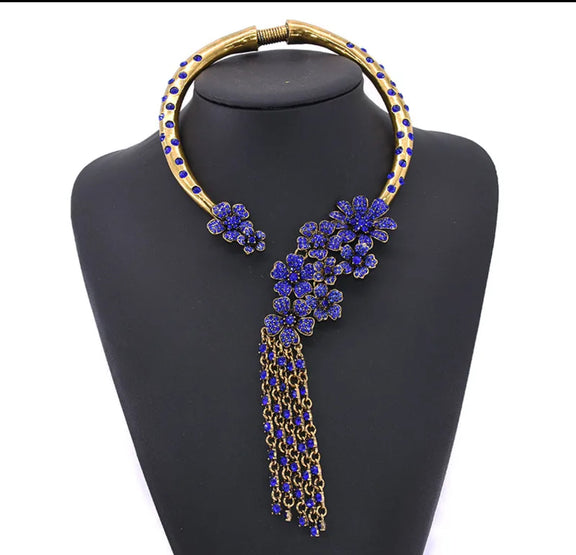 Bold vintage gold statement necklace featuring sapphire blue floral accents and cascading chain tassels, displayed on a black mannequin bust