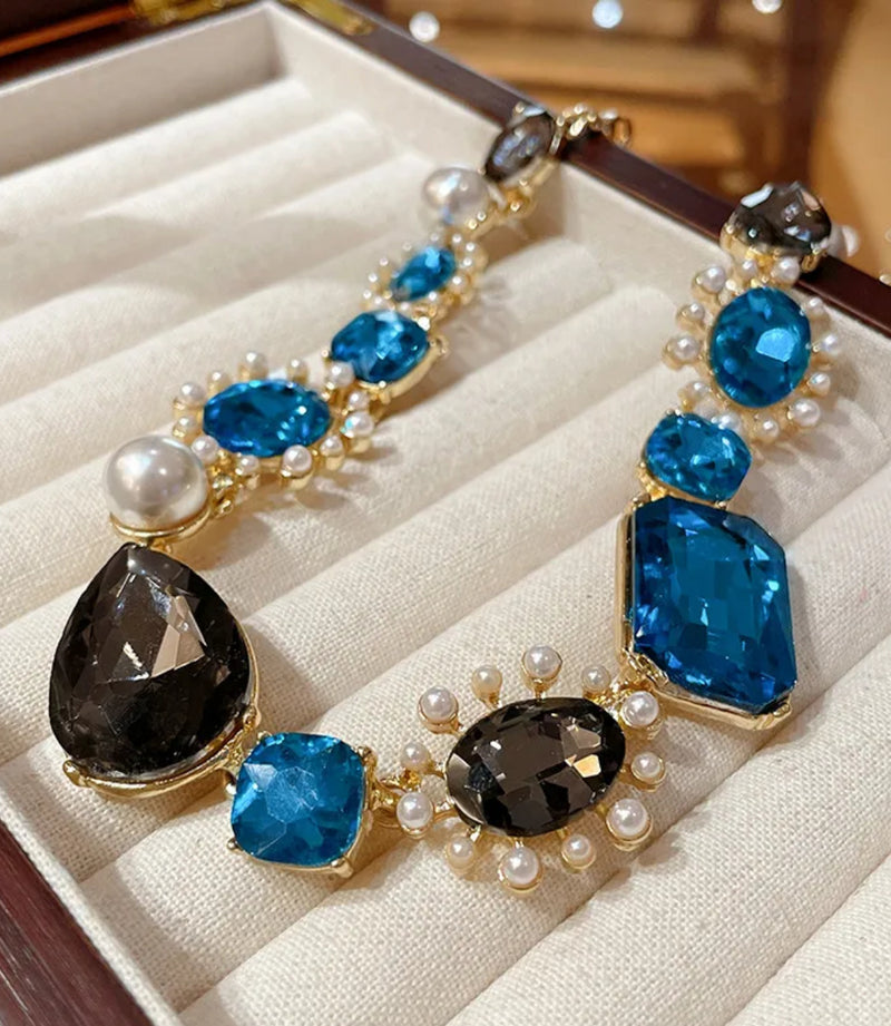 Bold gold-tone necklace with large sapphire-blue and smoky-gray gemstones, framed by pearls and arranged in an elegant, vintage-inspired design.
