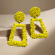 Bold yellow geometric statement earrings with an intricate floral texture, featuring a round stud top and a hollow rectangular drop design, displayed outdoors on a natural surface.”