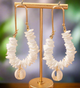 Gold hoop earrings with stacked white shell chips and a hanging cowrie shell, displayed on sand with a tropical backdrop.