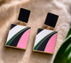 Zeta Mini earrings from Desiree W.A.R Store feature a modern geometric design with black, white, green, and pink sections on a gold border, displayed on a beige surface.