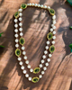 Long layered necklace featuring alternating white pearl beads and oval green gemstones framed in gold-tone settings, displayed on a black velvet bust. The necklace can be worn as a single long strand or wrapped into a double-layered style.