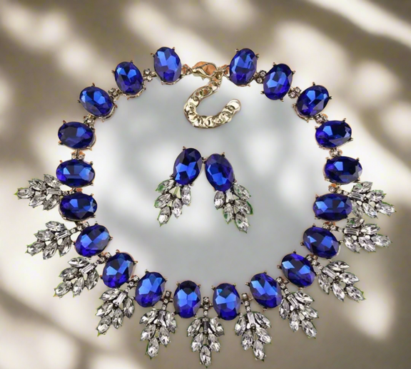 The Imani Lux Set from Desiree W.A.R Store features a geometric rhinestone necklace and short glass earrings set with large blue gemstones and leaf-shaped silver details, displayed on a neutral background.