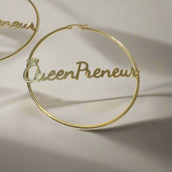 A pair of large, elegant gold hoop earrings featuring the word “QueenPreneur” in a bold cursive script, with a delicate crown accent over the “Q.” The earrings have a secure latch-back closure and a stylish, empowering design perfect for entrepreneurs and boss queens.
