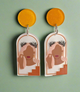 A pair of resin statement earrings featuring an abstract feminine silhouette framed within an arched design, accented with warm earthy tones and golden-yellow tops, hanging elegantly against a soft green background.