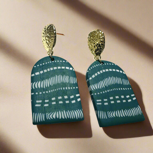 A pair of handcrafted statement earrings featuring gold-tone hammered metal studs with teal-green arched pendants adorned with white tribal patterns, designed for bold, elegant styling