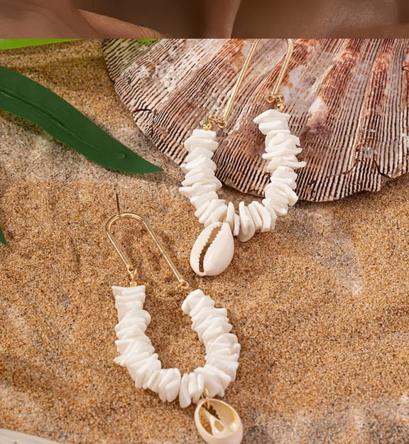 Gold hoop earrings with stacked white shell chips and a hanging cowrie shell, displayed on sand with a tropical backdrop.