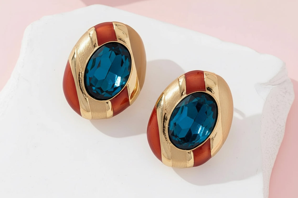 Gold and red earrings with blue gemstones on a white stand against a pink background