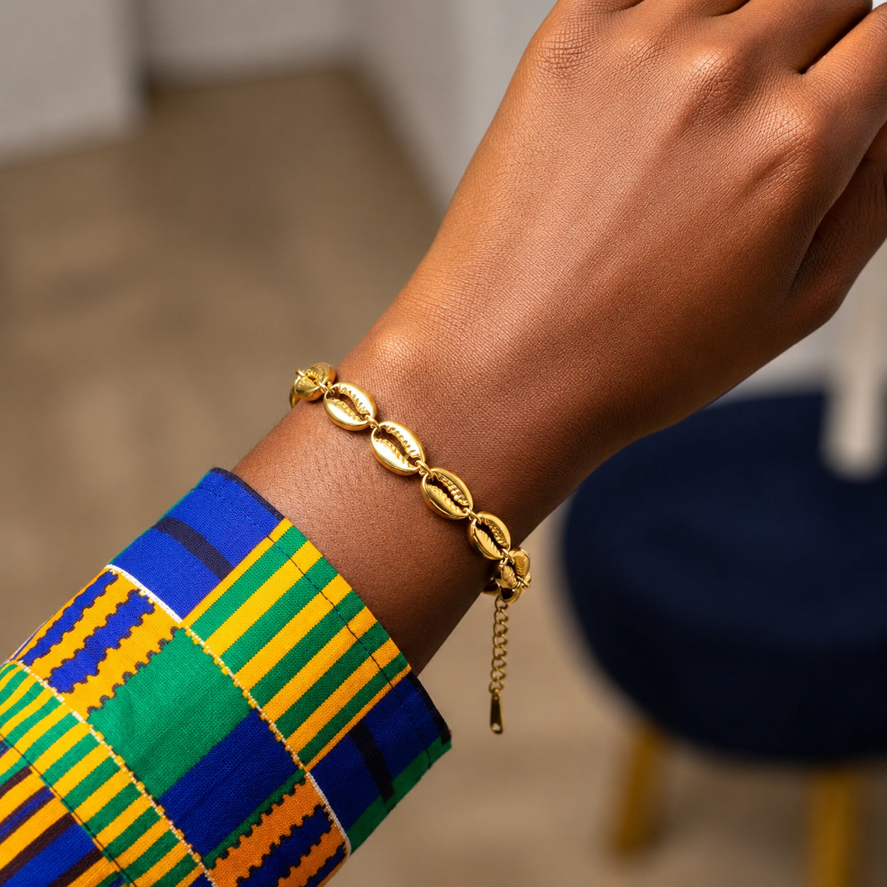 A gold chain bracelet with cowrie shell links worn on a person's wrist, paired with a colorful patterned cuff in blue, green, yellow, and orange.