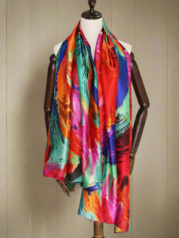 A luxurious silk scarf with a vibrant abstract design featuring bold red, blue, green, and orange hues. The fabric has a glossy finish, draping elegantly for a statement look.