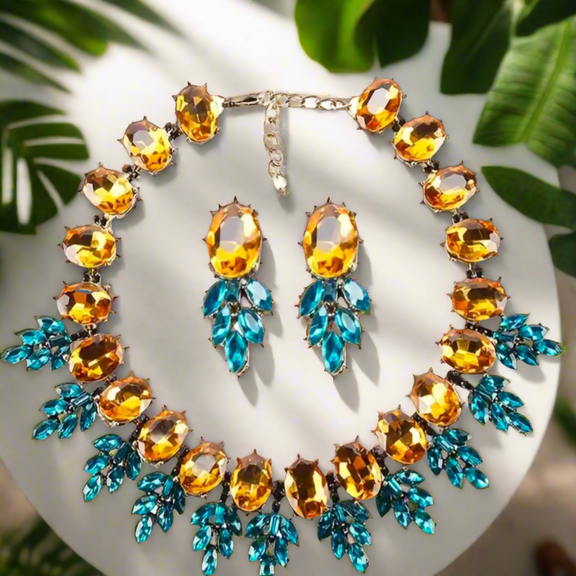The Imani Lux Set from Desiree W.A.R Store includes a Geometric Rhinestone Necklace with amber oval stones and blue leaf-shaped stones, paired with short glass earrings for an elegant and cohesive look.