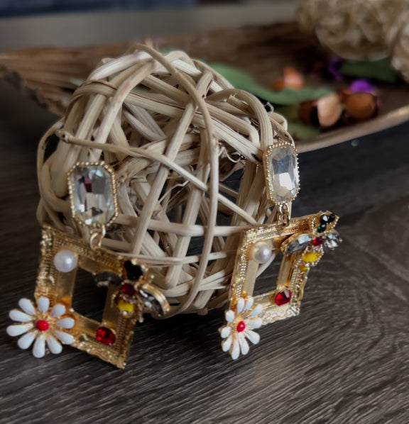 Close-up of gold rectangular drop earrings with rhinestone bee, white daisy, faux pearl, and crystal stud, placed against a rustic background.