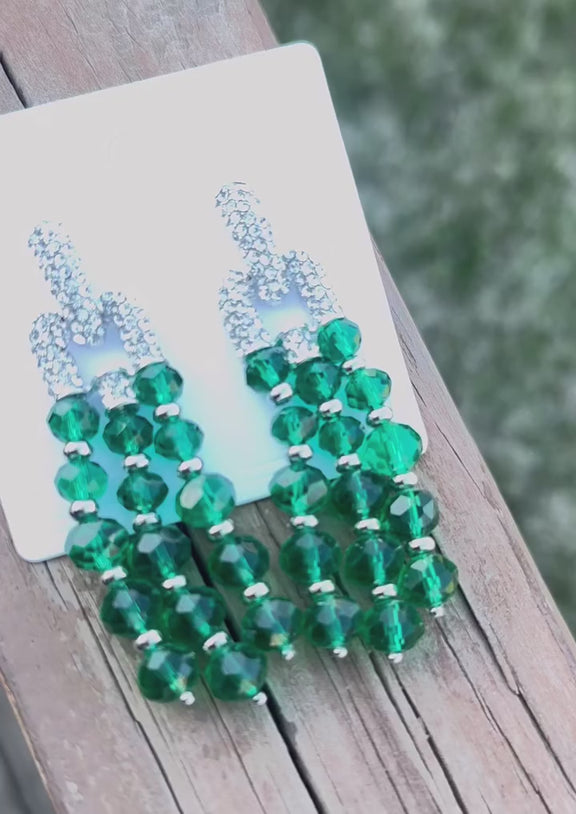 A pair of elegant drop earrings featuring cascading strands of faceted emerald green beads, accented with silver-toned spacers and topped with sparkling rhinestone-encrusted link connectors, displayed on a light surface with green palm leaves framing the image.