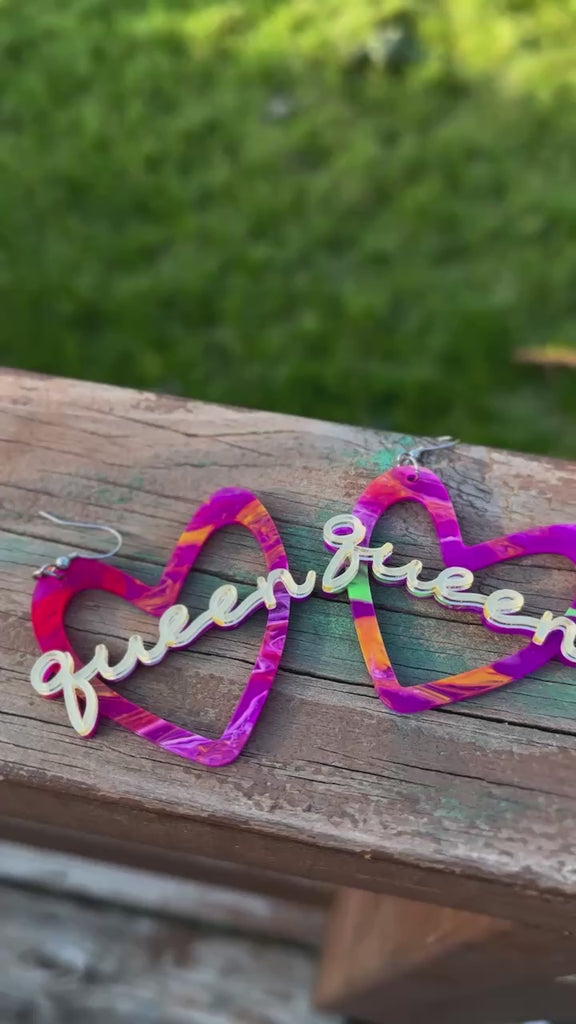 A pair of vibrant, multicolored heart-shaped earrings with the word “Queen” in gold-tone script across the center. The earrings have silver-tone hooks and feature a blend of purple, pink, orange, and green hues.