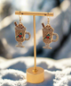 Holiday Cocoa Sparkle Earrings shaped like festive hot chocolate mugs with whipped cream, cinnamon sticks, and colorful crystal accents in gold, red, and green.