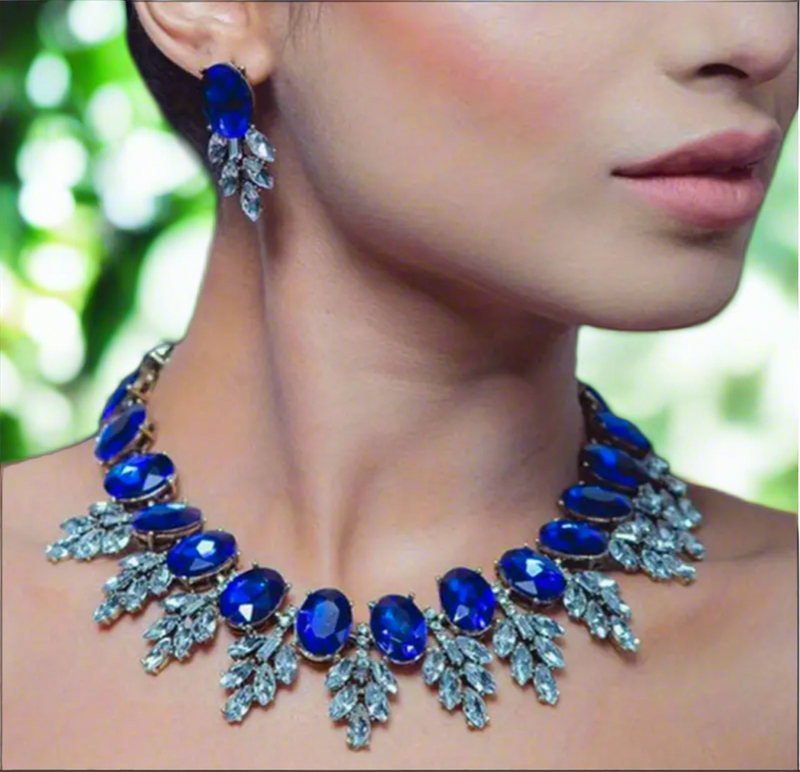 A woman showcases the Imani Lux Set from Desiree W.A.R Store, featuring short glass earrings and a necklace with blue gemstones and geometric rhinestone leaf designs, set against a blurred green background.