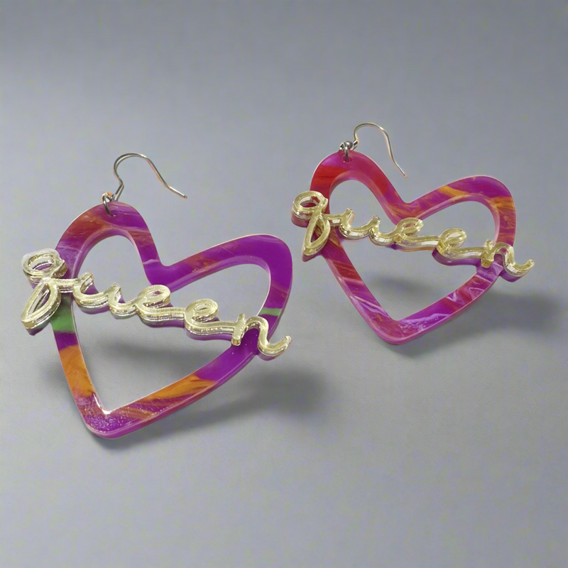 A pair of vibrant, multicolored heart-shaped earrings with the word “Queen” in gold-tone script across the center. The earrings have silver-tone hooks and feature a blend of purple, pink, orange, and green hues.