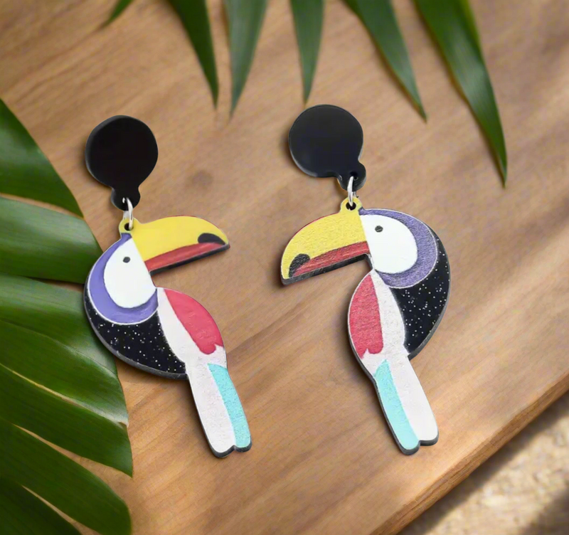 Colorful toucan-shaped statement earrings with yellow beaks, red and blue accents, and black circular tops, hanging on a display rod.
