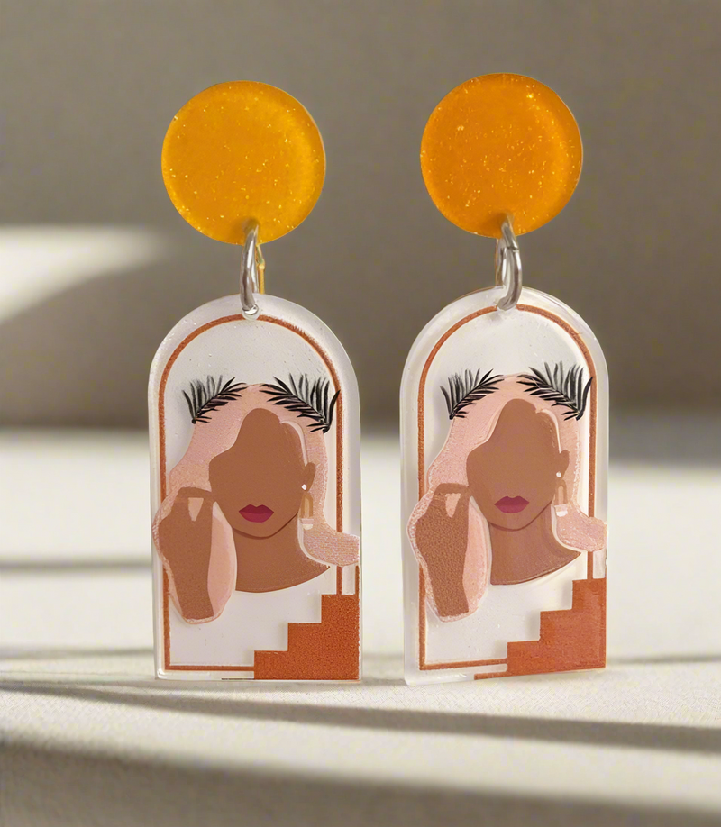 A pair of resin statement earrings featuring an abstract feminine silhouette framed within an arched design, accented with warm earthy tones and golden-yellow tops, hanging elegantly against a soft green background.