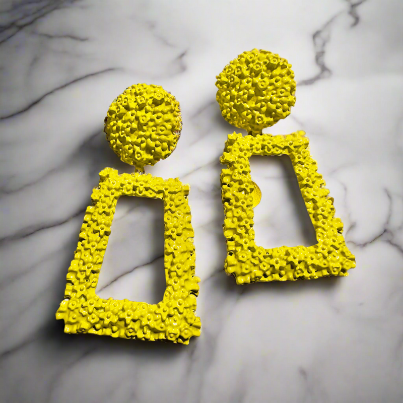 Bold yellow geometric statement earrings with an intricate floral texture, featuring a round stud top and a hollow rectangular drop design, displayed outdoors on a natural surface.”
