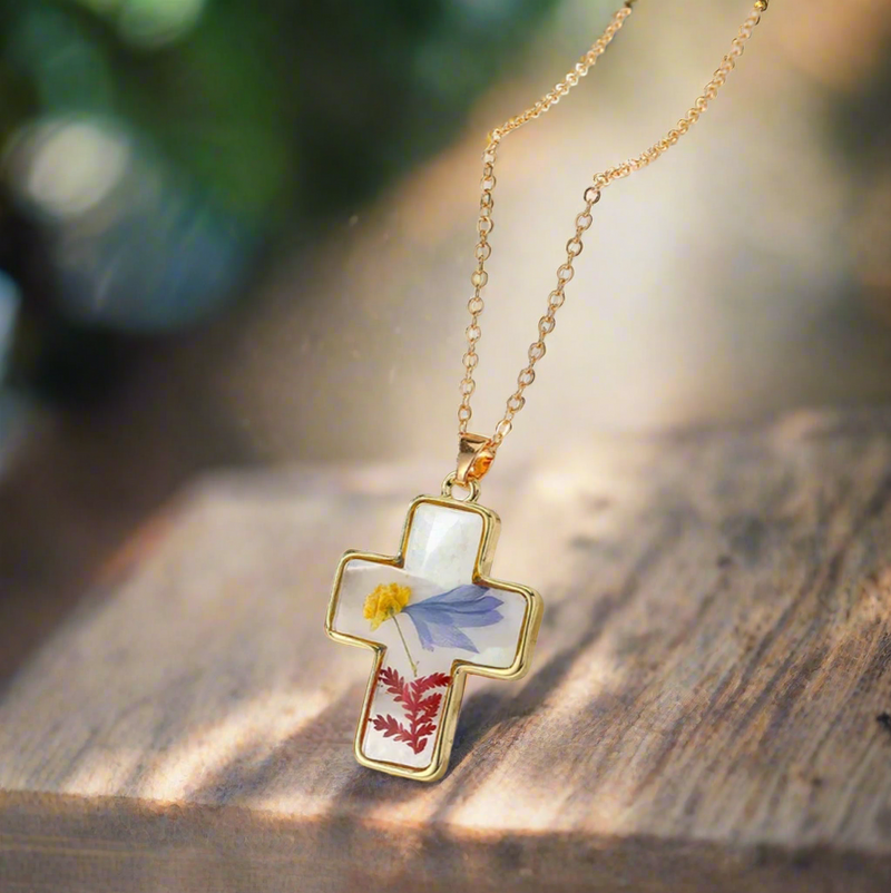 Gold-outlined cross pendant necklace featuring real pressed yellow, blue, and red flowers encased in clear resin, displayed on a delicate gold chain against a white textured background.
