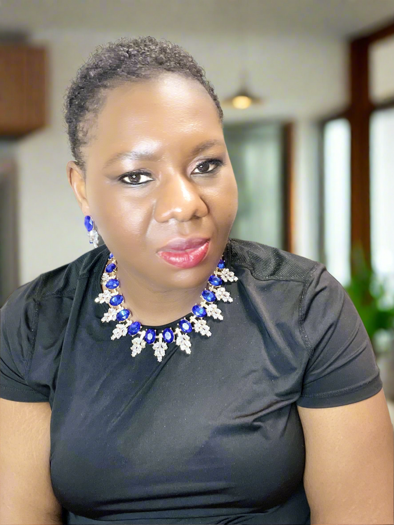 A person in a black shirt sports the Imani Lux Set from Desiree W.A.R Store, featuring chic short glass earrings and a geometric rhinestone necklace, while seated indoors.