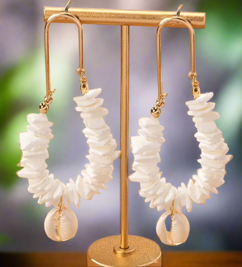 Gold hoop earrings with stacked white shell chips and a hanging cowrie shell, displayed on sand with a tropical backdrop.