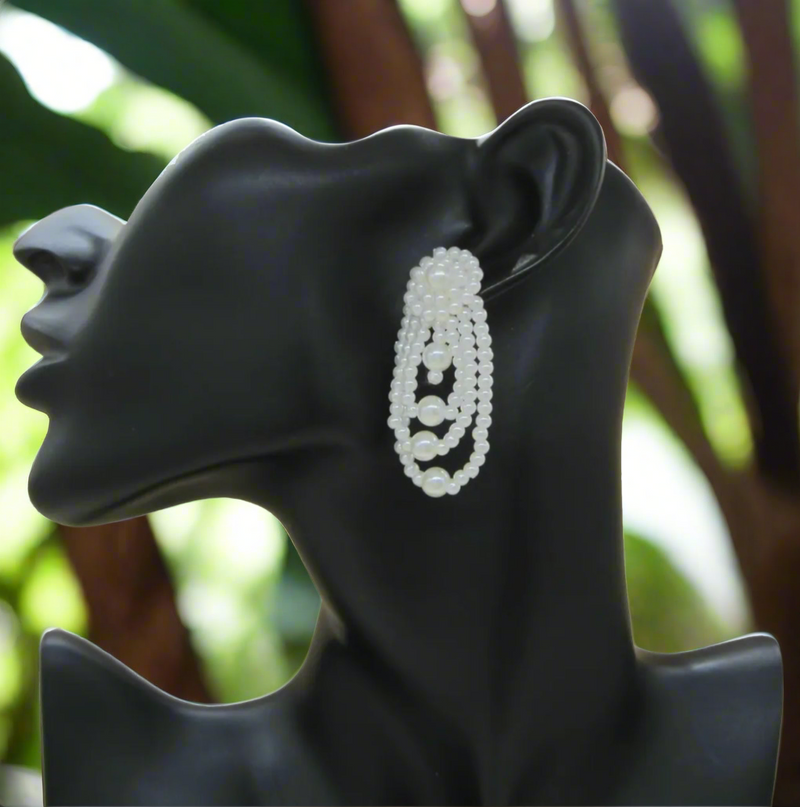 A pair of cascading pearl statement earrings featuring layered oval loops of ivory faux pearls with a clustered pearl stud, displayed on a wooden surface
