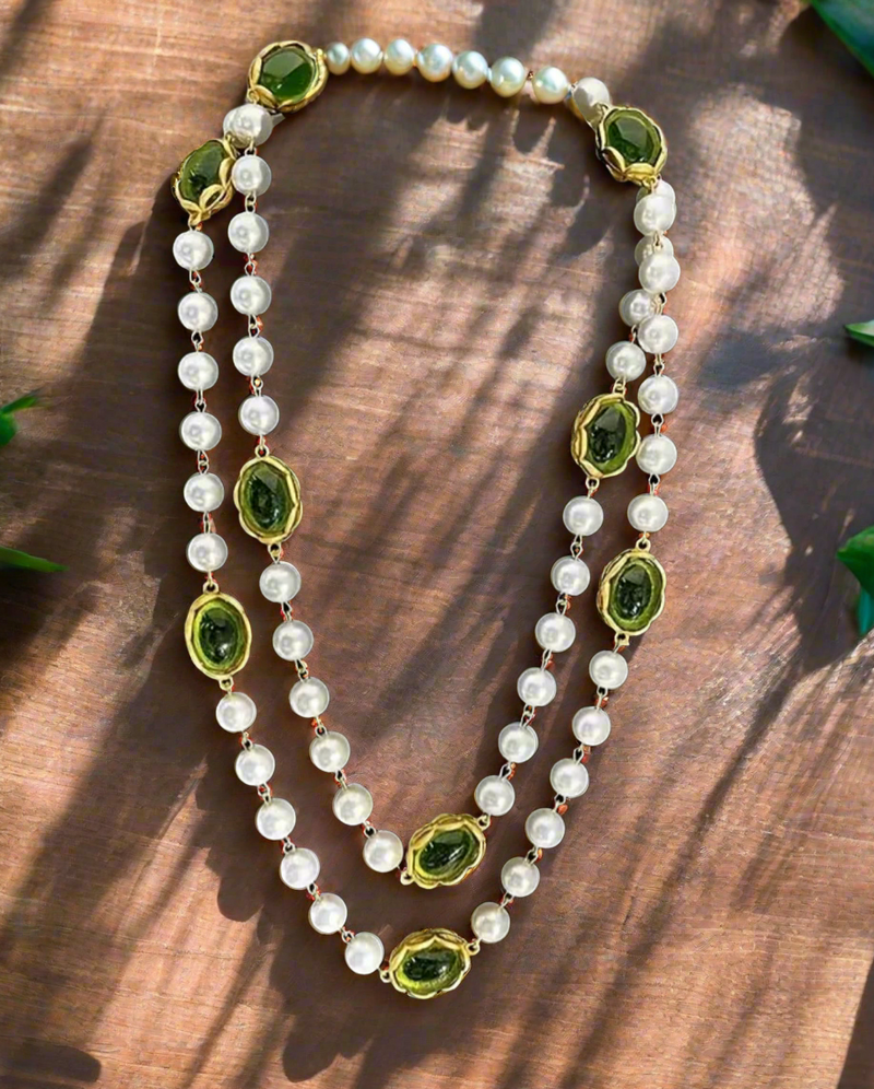Long layered necklace featuring alternating white pearl beads and oval green gemstones framed in gold-tone settings, displayed on a black velvet bust. The necklace can be worn as a single long strand or wrapped into a double-layered style.

