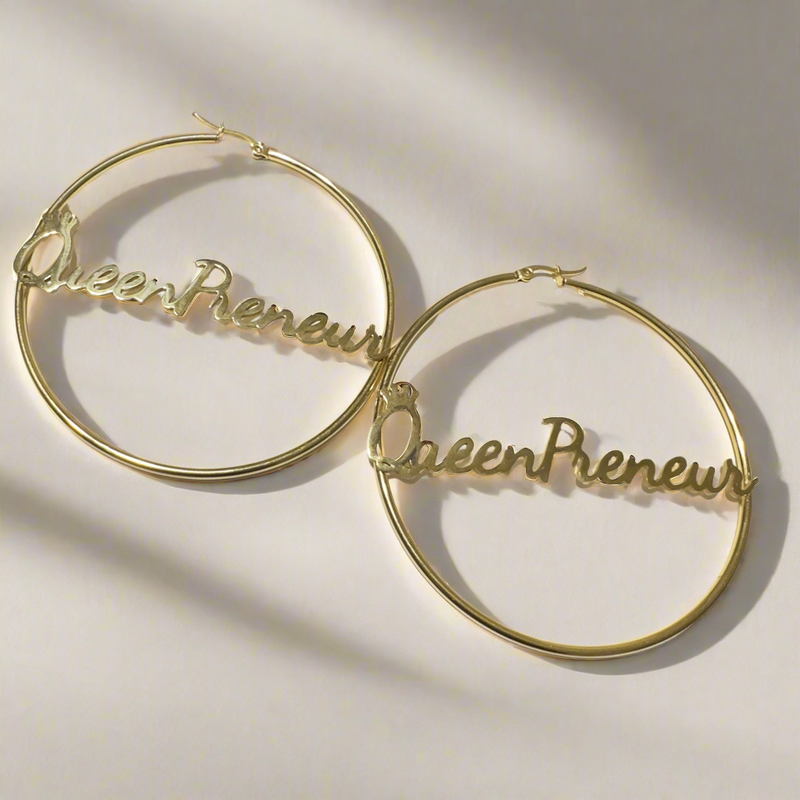 A pair of large, elegant gold hoop earrings featuring the word “QueenPreneur” in a bold cursive script, with a delicate crown accent over the “Q.” The earrings have a secure latch-back closure and a stylish, empowering design perfect for entrepreneurs and boss queens.