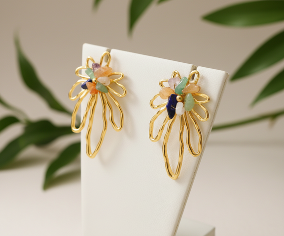 Gold floral earrings with colorful stones on a white stand with green leaves in the background