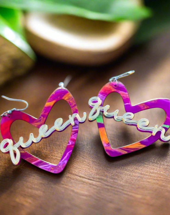 A pair of vibrant, multicolored heart-shaped earrings with the word “Queen” in gold-tone script across the center. The earrings have silver-tone hooks and feature a blend of purple, pink, orange, and green hues.