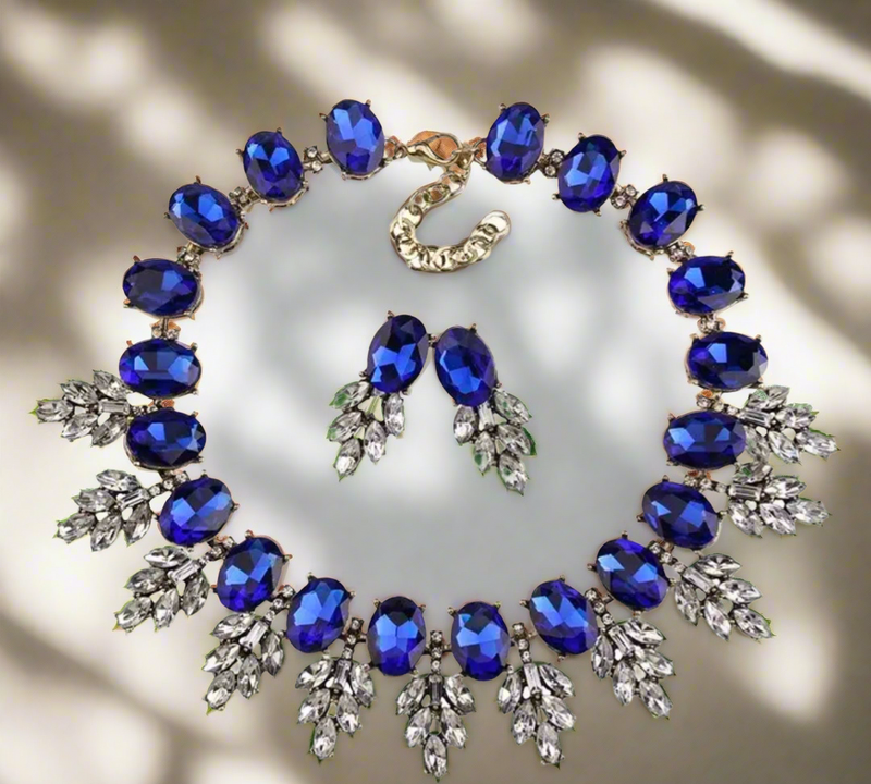 The Imani Lux Set from Desiree W.A.R Store features a geometric rhinestone necklace and short glass earrings set with large blue gemstones and leaf-shaped silver details, displayed on a neutral background.