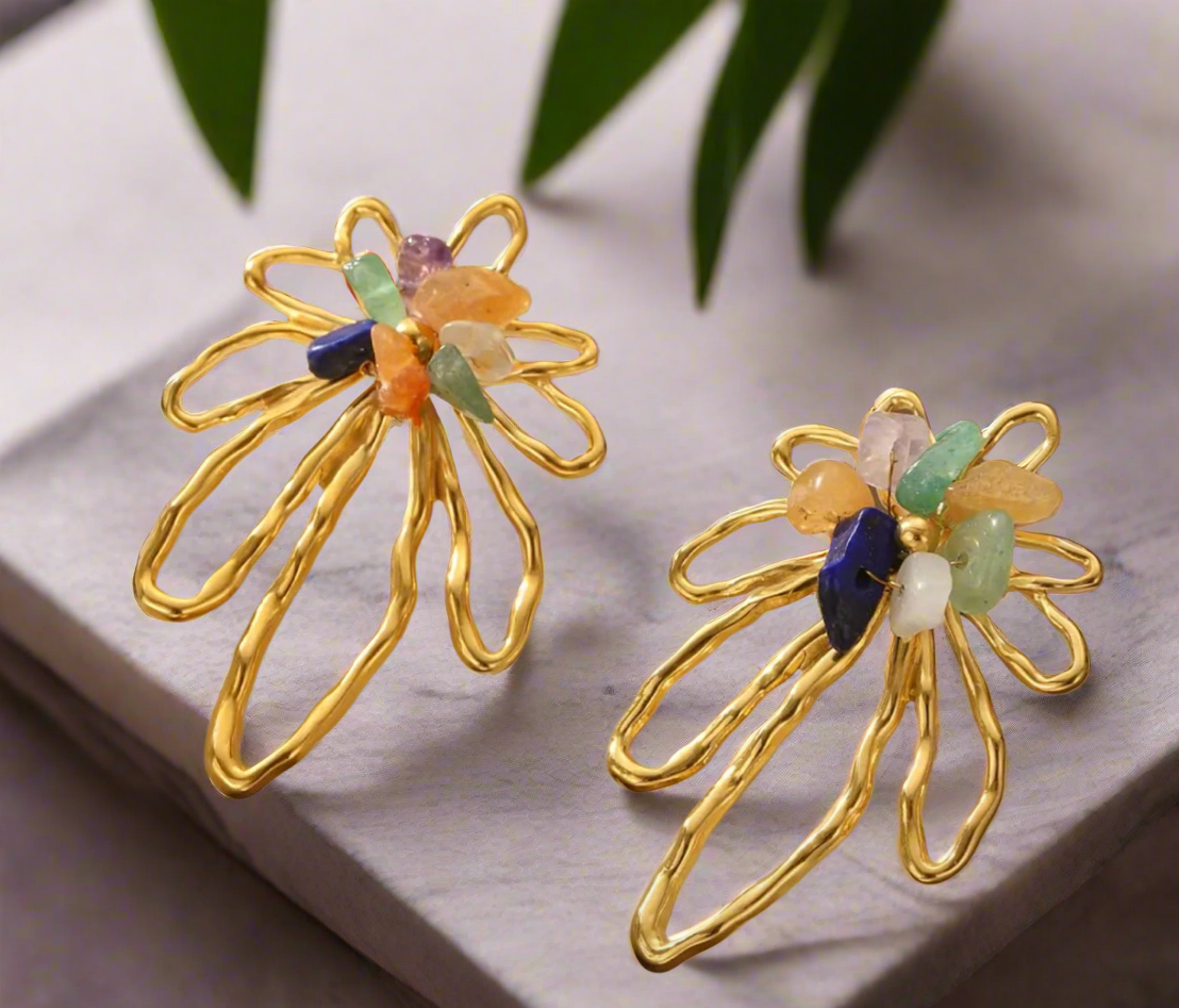 A pair of gold floral statement earrings with open petal design, accented by colorful natural gemstones including green, blue, orange, and purple tones, displayed on a neutral backgroun