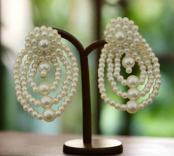 A pair of cascading pearl statement earrings featuring layered oval loops of ivory faux pearls with a clustered pearl stud, displayed on a wooden surface