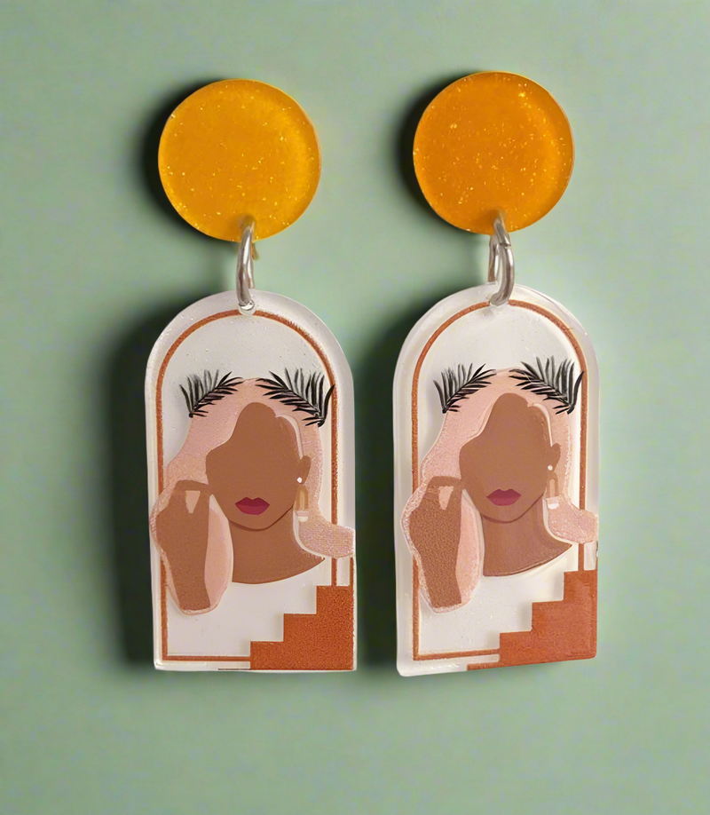 A pair of resin statement earrings featuring an abstract feminine silhouette framed within an arched design, accented with warm earthy tones and golden-yellow tops, hanging elegantly against a soft green background.