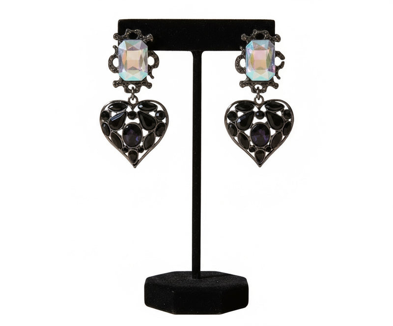 Black heart-shaped statement earrings with iridescent crystal tops, placed on a neutral stone background. Elegant jewelry from Women Are Rubies.
