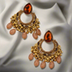 A pair of gold-tone chandbali earrings with a large amber teardrop stone at the top, decorated with golden beads, leaf accents, and dangling oval beads, placed on a wooden background.