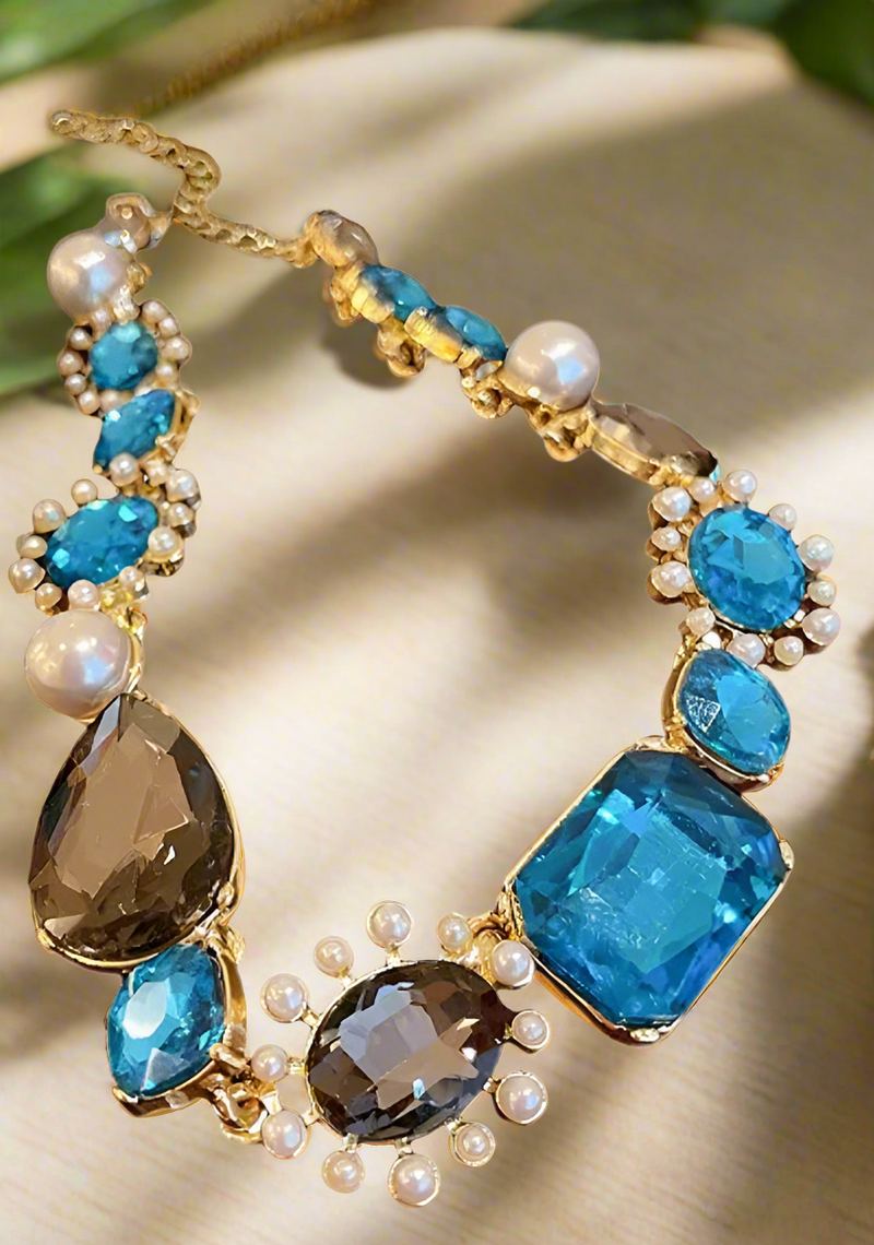 Bold gold-tone necklace with large sapphire-blue and smoky-gray gemstones, framed by pearls and arranged in an elegant, vintage-inspired design.
