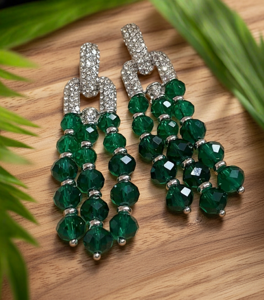 A pair of elegant drop earrings featuring cascading strands of faceted emerald green beads, accented with silver-toned spacers and topped with sparkling rhinestone-encrusted link connectors, displayed on a light surface with green palm leaves framing the image.