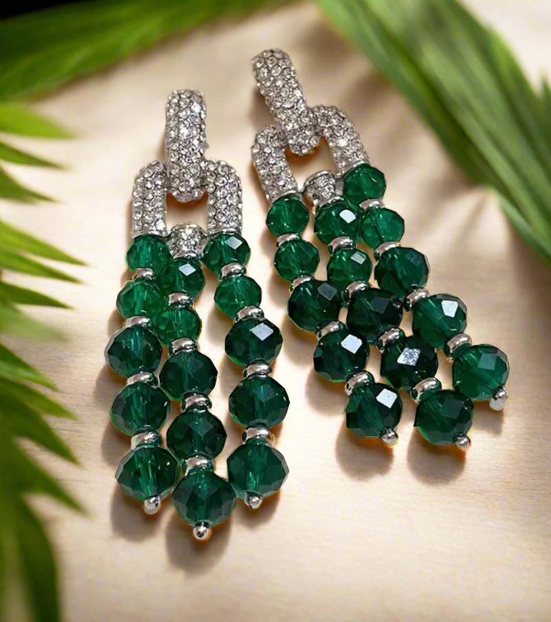 A pair of elegant drop earrings featuring cascading strands of faceted emerald green beads, accented with silver-toned spacers and topped with sparkling rhinestone-encrusted link connectors, displayed on a light surface with green palm leaves framing the image.