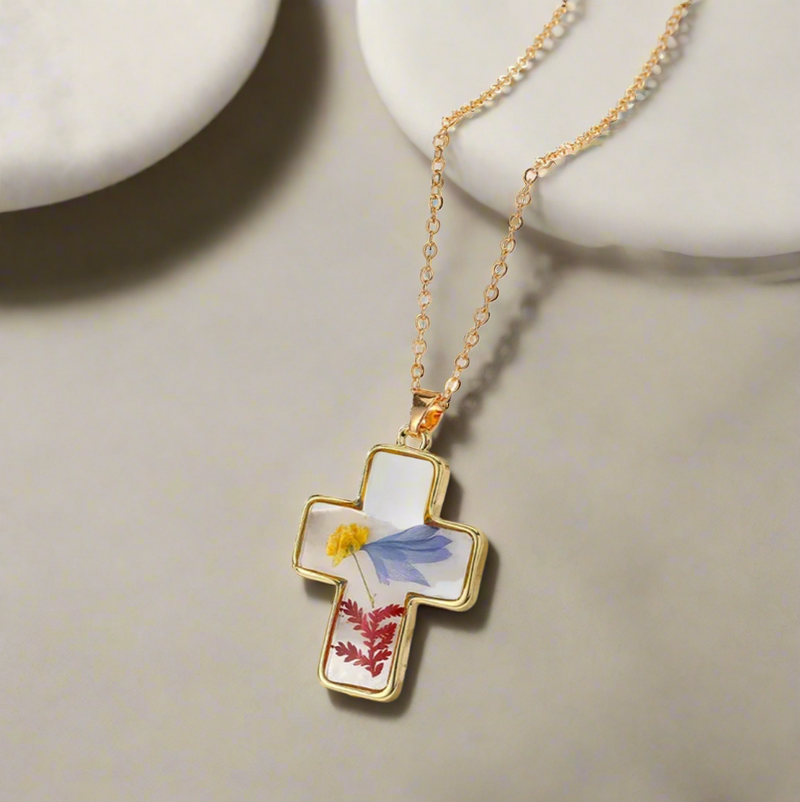 Gold-outlined cross pendant necklace featuring real pressed yellow, blue, and red flowers encased in clear resin, displayed on a delicate gold chain against a white textured background.
