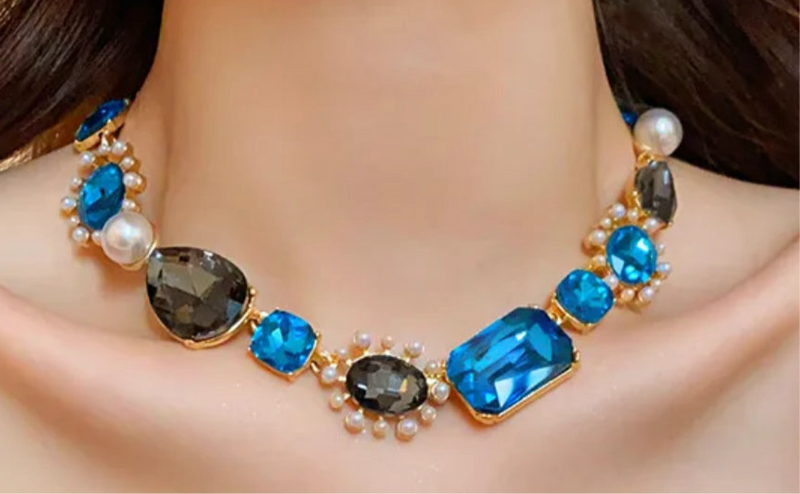 Bold gold-tone necklace with large sapphire-blue and smoky-gray gemstones, framed by pearls and arranged in an elegant, vintage-inspired design.
