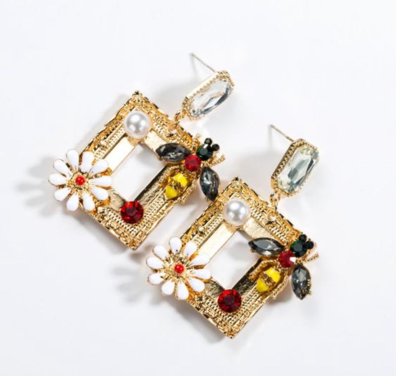 Close-up of gold rectangular drop earrings with rhinestone bee, white daisy, faux pearl, and crystal stud, placed against a rustic background.
