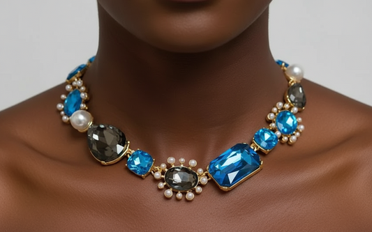 Bold gold-tone necklace with large sapphire-blue and smoky-gray gemstones, framed by pearls and arranged in an elegant, vintage-inspired design.
