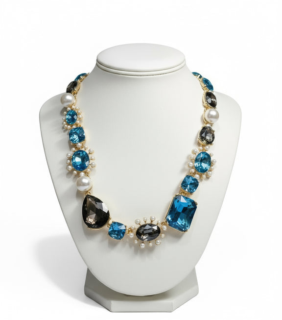 Bold gold-tone necklace with large sapphire-blue and smoky-gray gemstones, framed by pearls and arranged in an elegant, vintage-inspired design.
