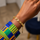 A gold chain bracelet with cowrie shell links worn on a person's wrist, paired with a colorful patterned cuff in blue, green, yellow, and orange.