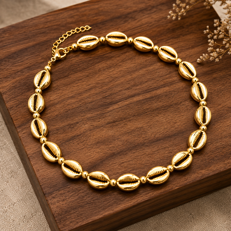 A gold-toned beaded necklace with cowrie shell-shaped beads arranged in a circle on a dark wooden block, with dried floral stems in the corner.