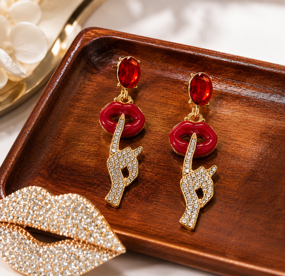 A pair of gold-tone drop earrings with red oval stones, red lip-shaped rings, and pavé-set crystal hand charms pointing upward, displayed on a dark wooden tray.